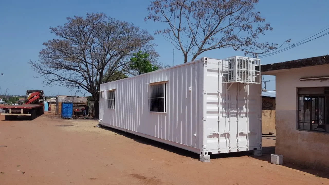 Modulo de contenedor blanco tipo oficina de obra con ventanas con rejas y equipo de aire, sobre blocks en un predio.