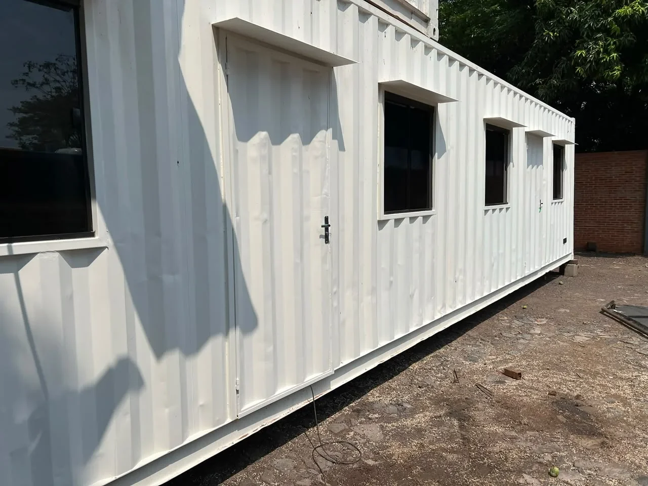 Modulo de oficina en contenedor blanco con puerta y ventanas de marco negro, sobre predio pavimentado.