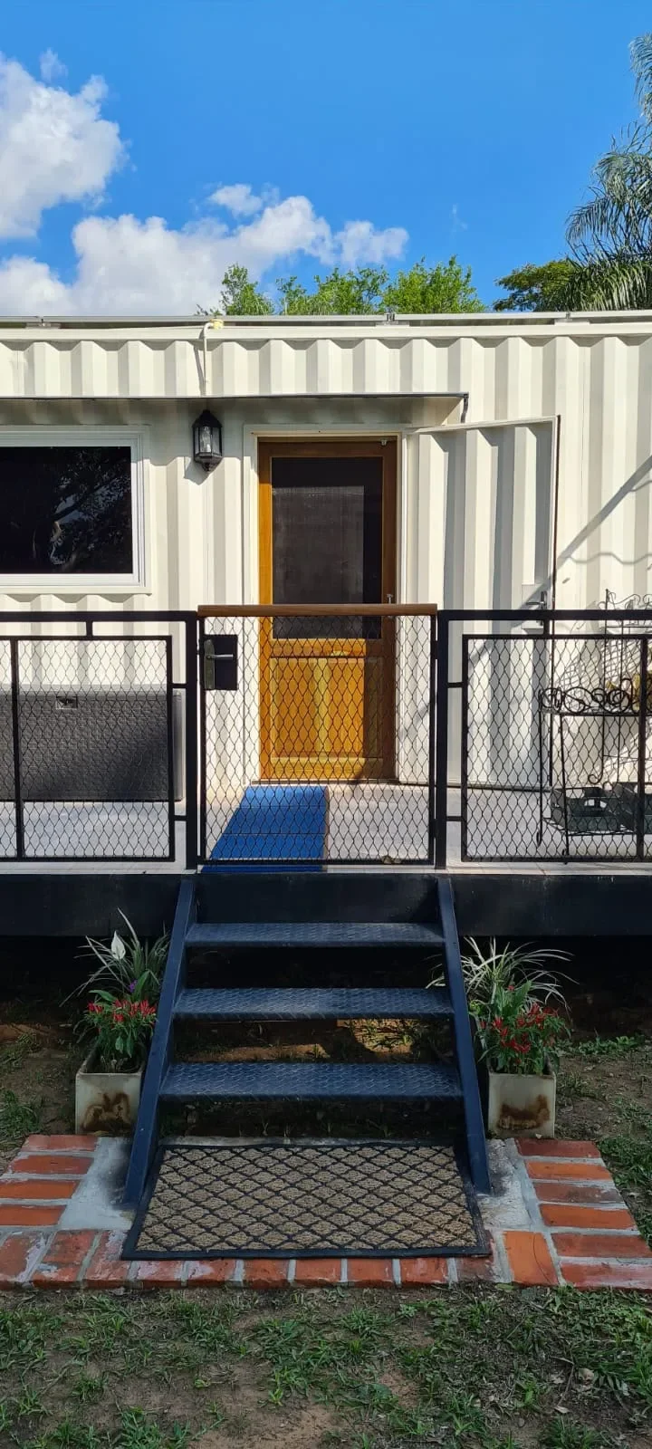 Entrada de cabana de contenedor blanca con puerta de madera, escalera de hormigon pintada de azul y barandas de hierro negro.