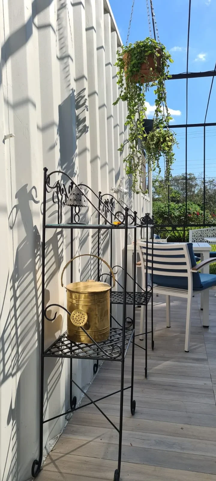 Detalle del deck de la cabana blanca: estanteria de hierro forjado con regadera dorada, planta colgante y sillones azules.