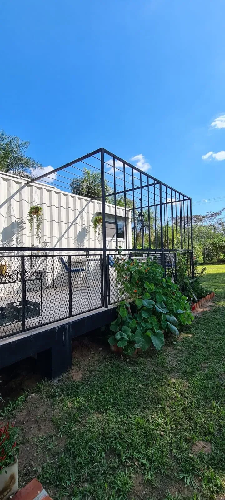 Deck con pergola de hierro negro junto a cabana de contenedor blanca, plantas y cesped.