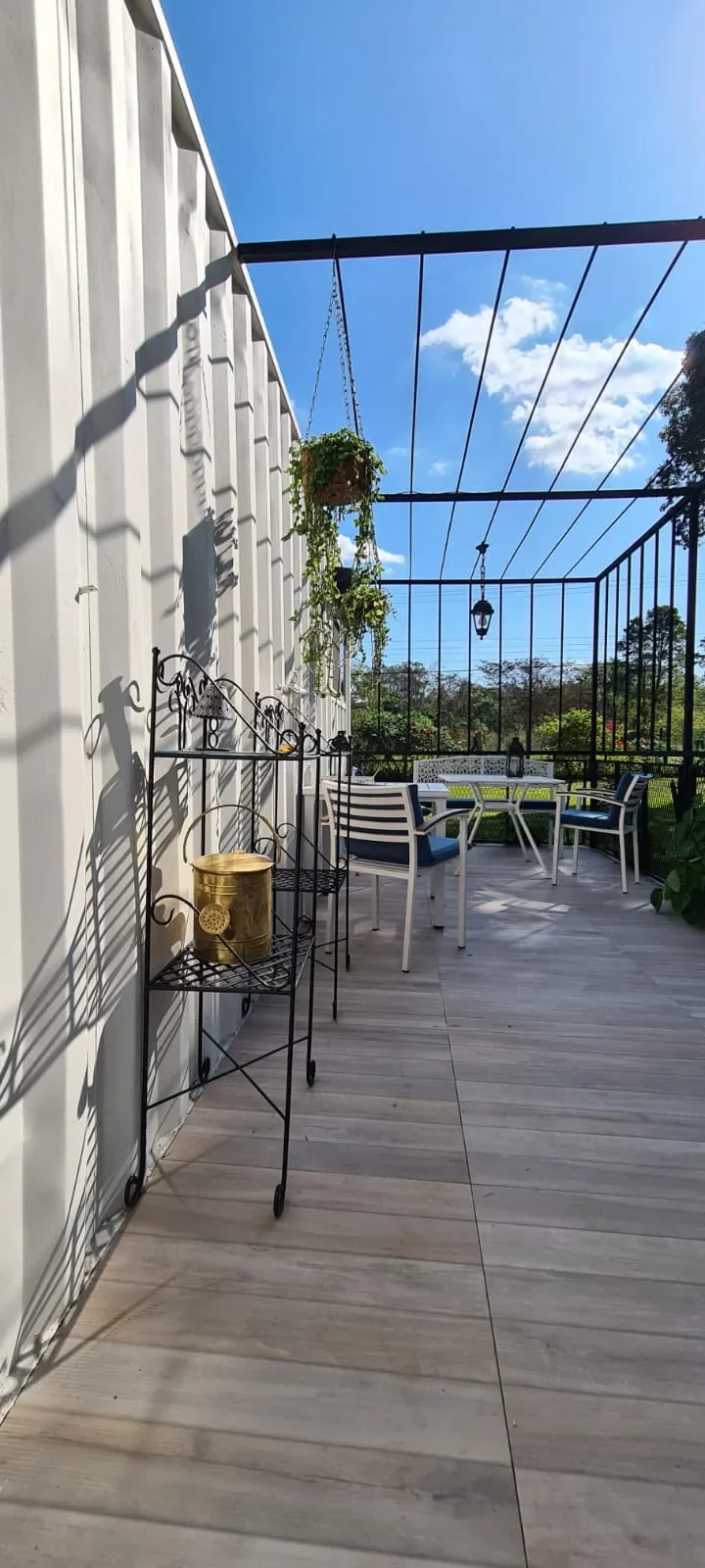 Deck con pergola de hierro negro de la cabana blanca, comedor de exterior con sillas de cojin azul y vista al jardin florido.