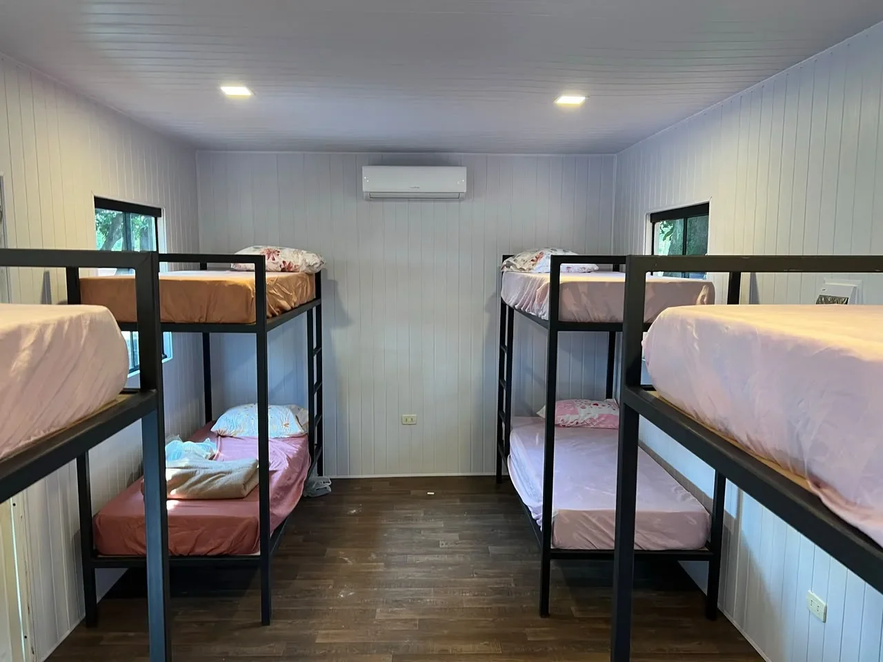 Dormitorio en contenedor con cuatro literas de hierro negro y ropa de cama rosa, paredes de machimbre blanco.