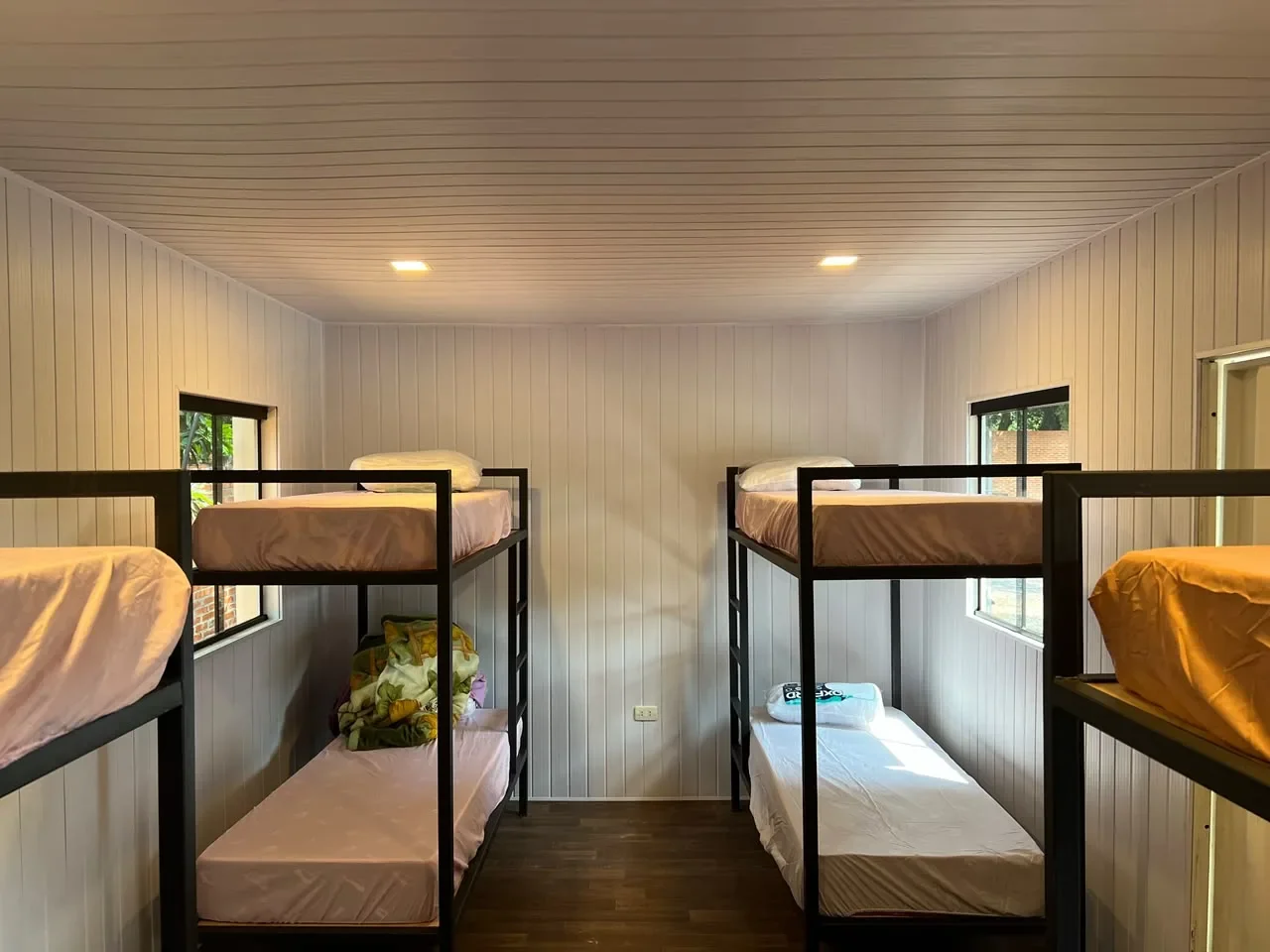 Dormitorio en contenedor con cuatro literas de hierro negro, paredes y cielorraso de machimbre blanco y piso de madera, tipo campamento.