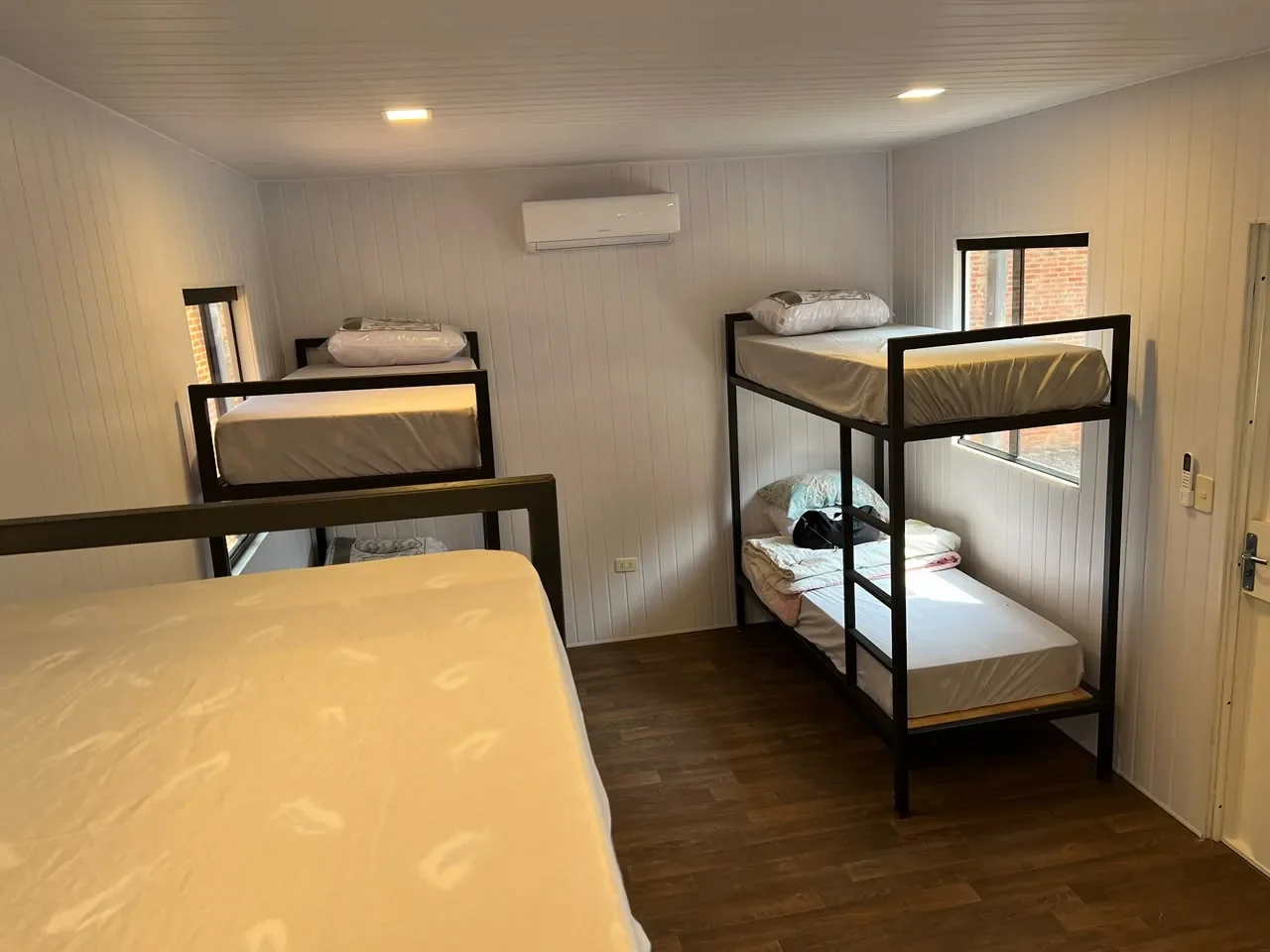 Dormitorio en contenedor con literas de hierro negro y una cama doble, paredes de machimbre blanco y piso de madera, uso campamento.