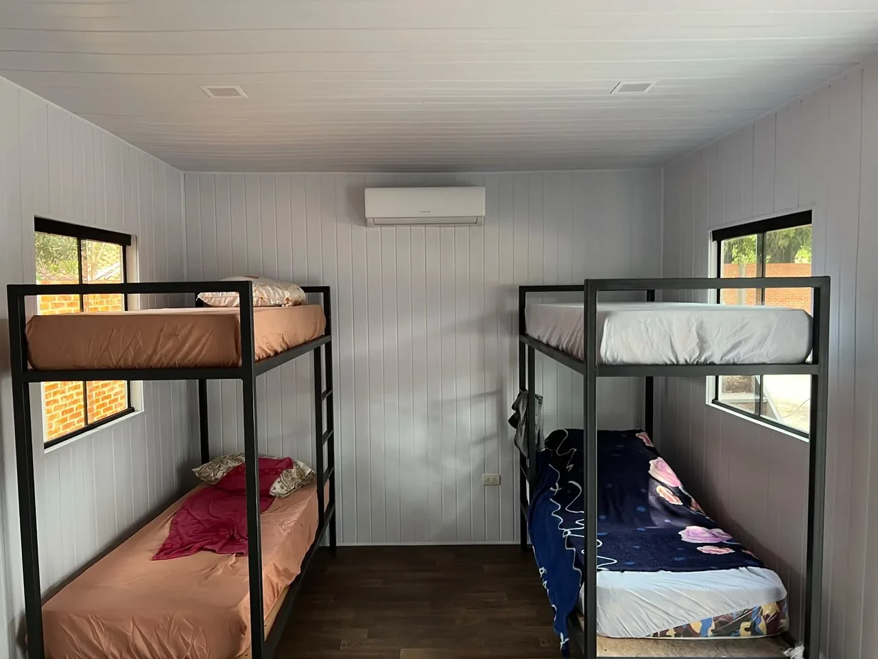 Dormitorio en contenedor con literas de hierro negro, paredes de machimbre blanco y piso de madera, tipo campamento.