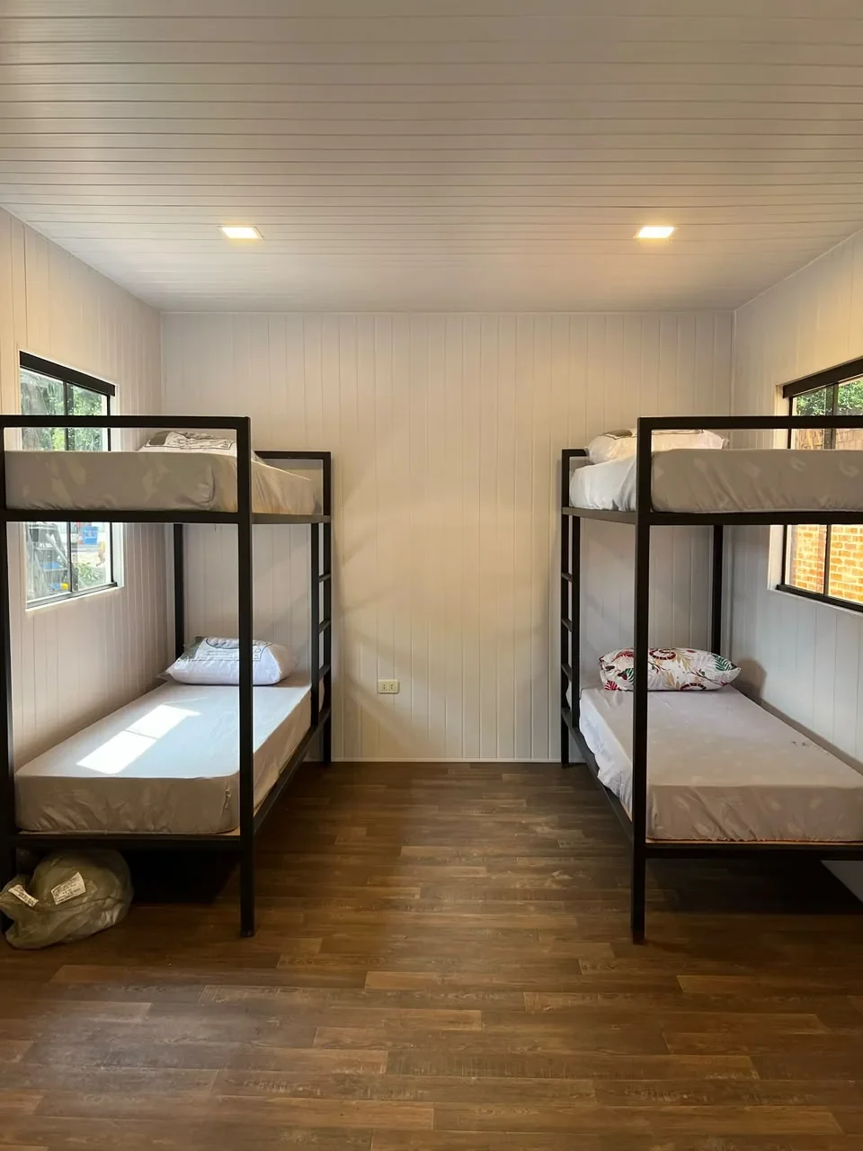 Dormitorio en contenedor con literas de hierro negro, paredes de machimbre blanco y piso de madera oscura.