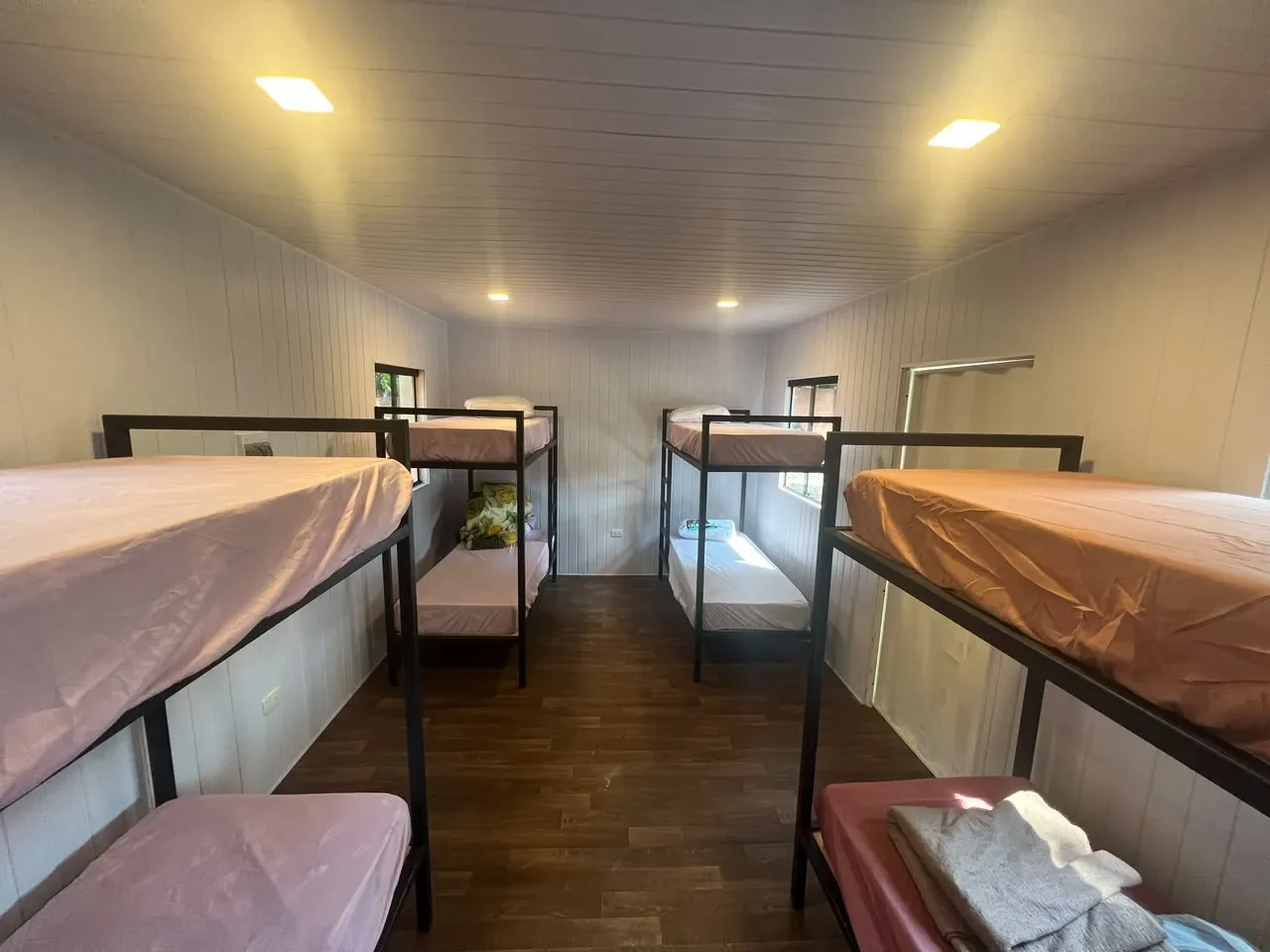 Dormitorio en contenedor con seis literas de hierro negro y ropa de cama rosa, paredes de machimbre blanco y piso de madera.