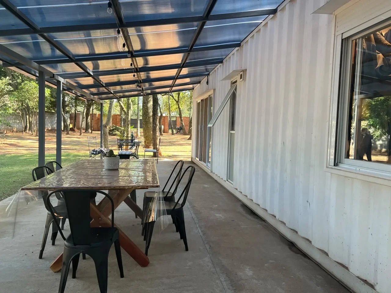 Galeria de casa de contenedor blanca con pergola plana, comedor rustico de madera y sillas negras sobre piso de hormigon, entorno arbolado.