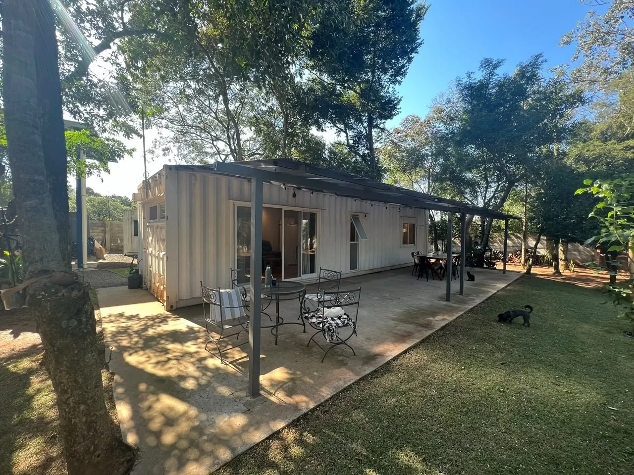 Casa de contenedor blanca de una planta con galeria techada, mobiliario de exterior y patio bajo arboles.