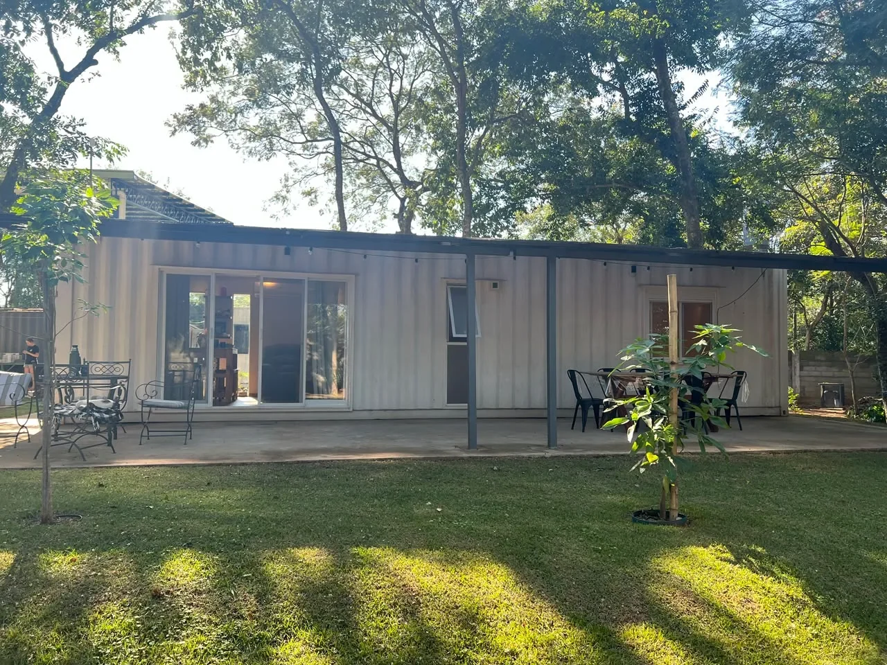 Casa de contenedor maritimo blanca de una planta con galeria techada, mesa y sillas de exterior, en terreno arbolado. Paraguay.