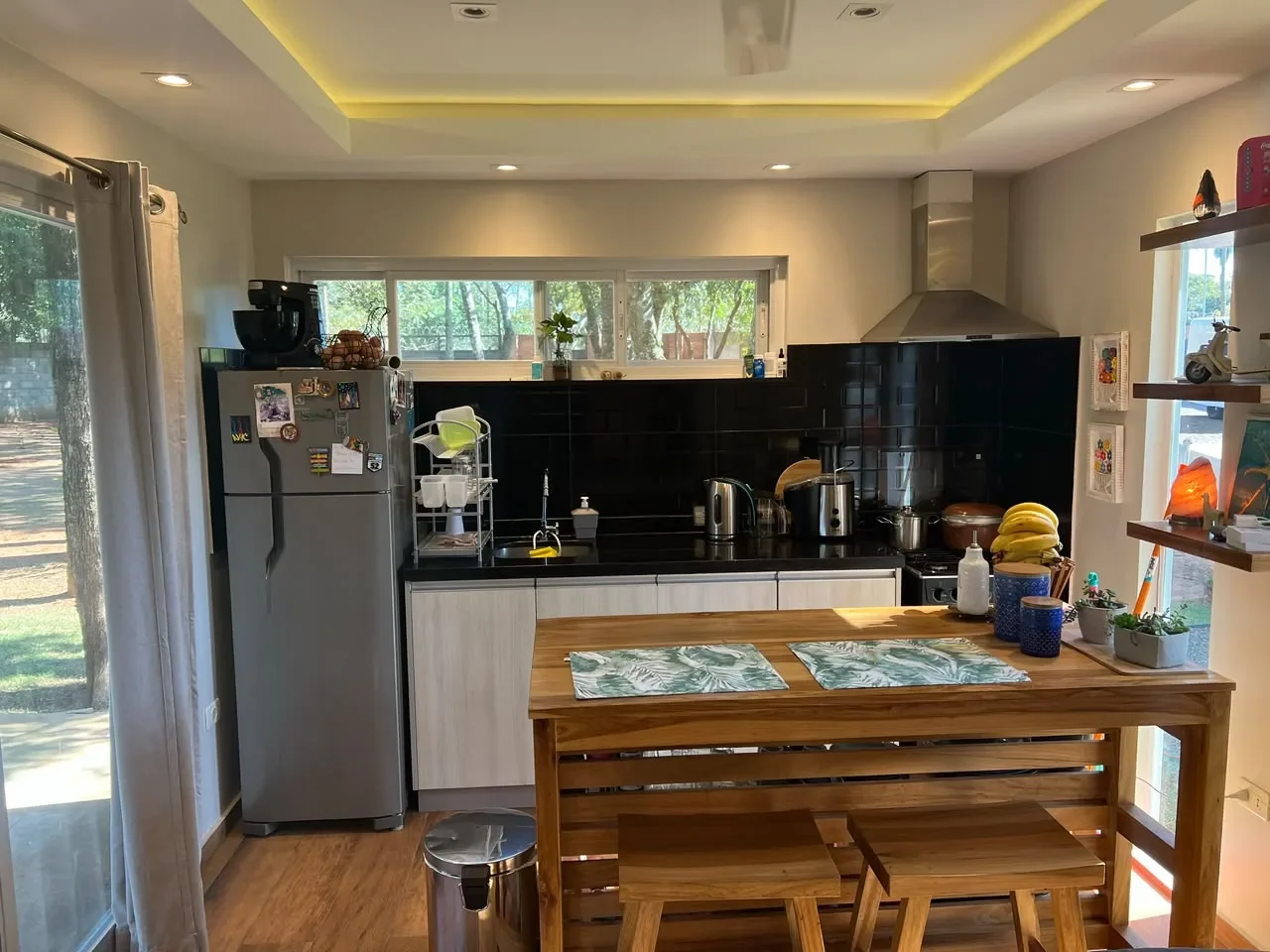 Cocina-comedor de casa de contenedor habitada con muebles claros, granito negro, salpicadero subway negro, mesa de madera con bancos y ventana al jardin.