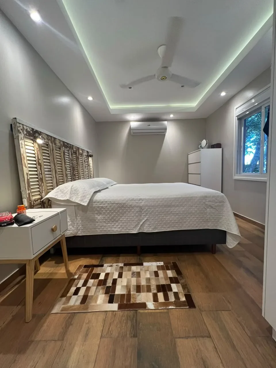 Dormitorio principal de casa de contenedor con cabecera de persianas recicladas, alfombra de cuero patchwork, cove LED y piso de madera.