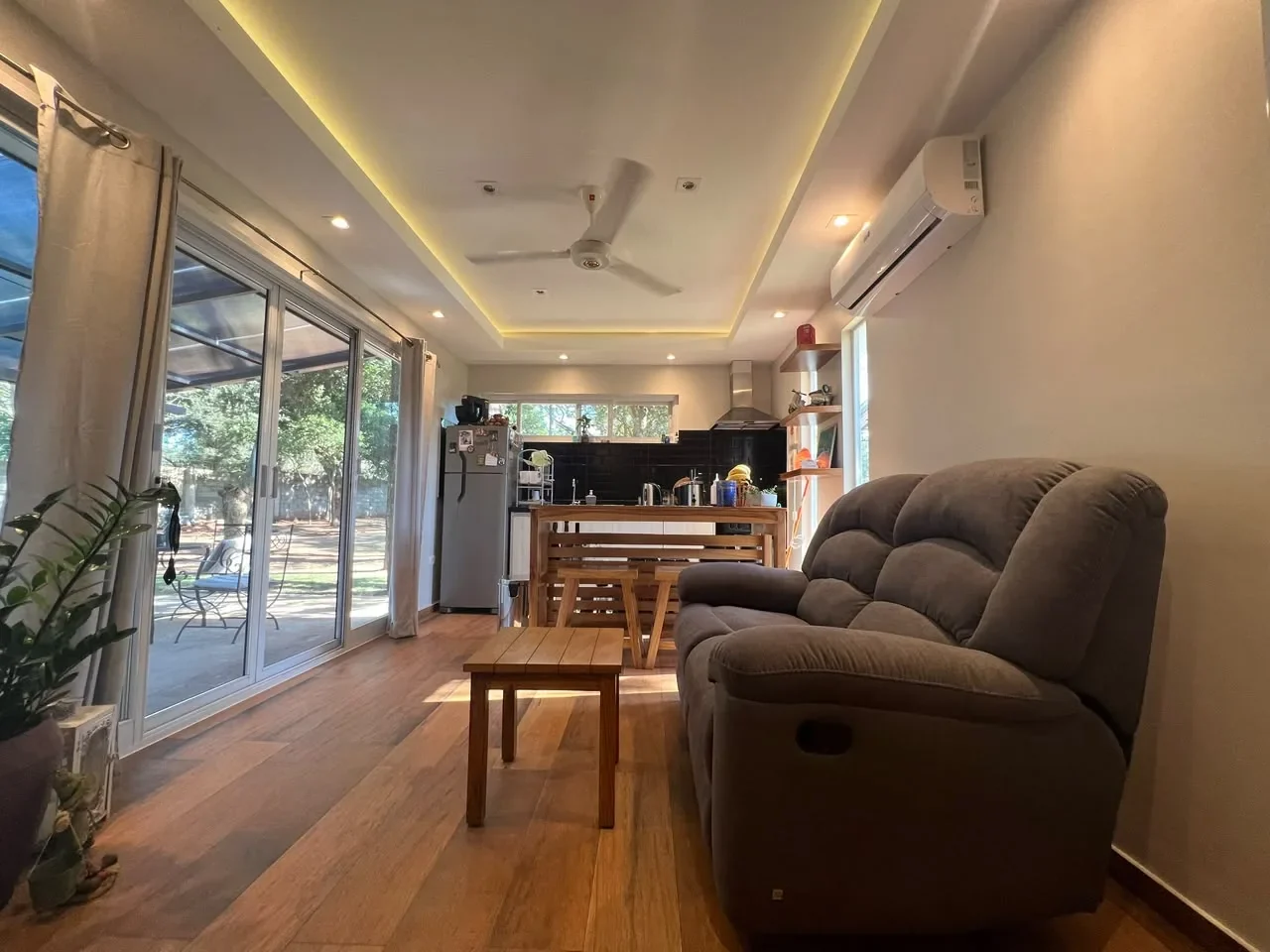 Living de casa de contenedor con sofa gris reclinable, cielorraso cove LED, cocina de subway negro al fondo y ventanal corredizo al jardin.