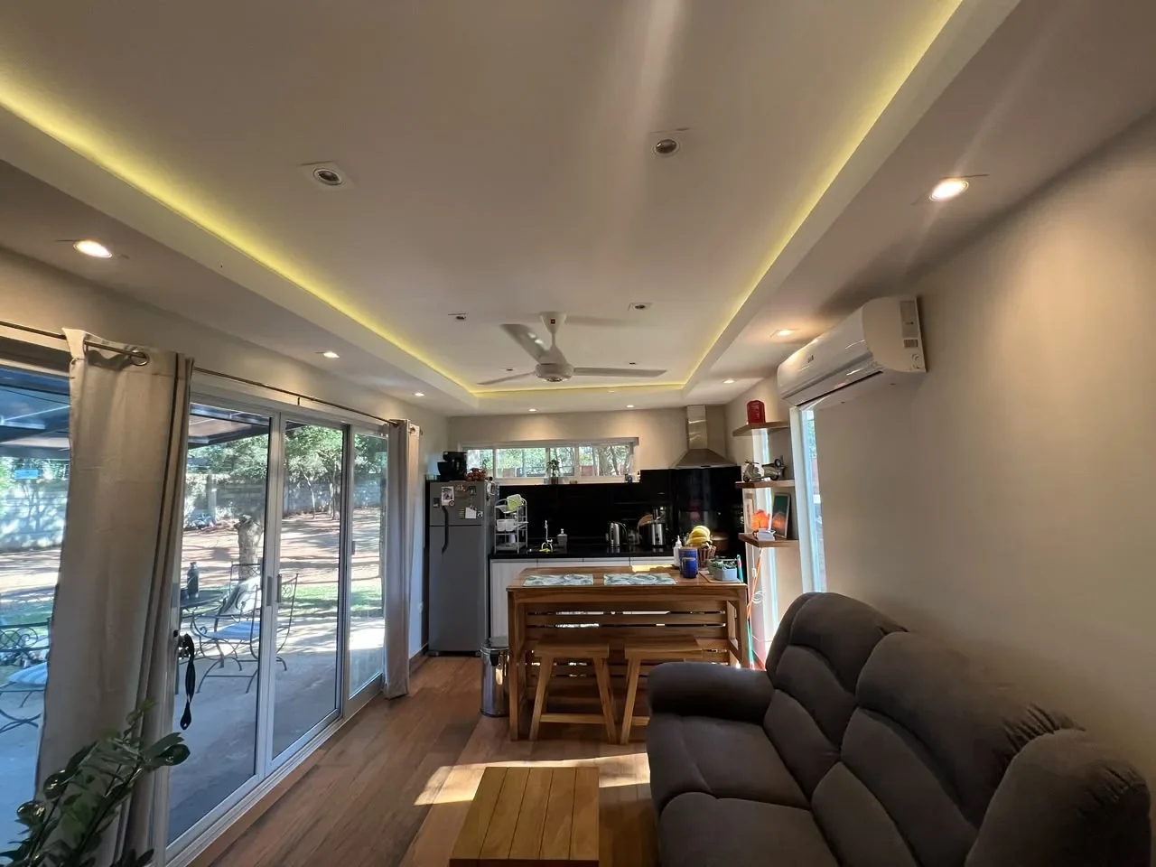 Living-cocina integrado de casa de contenedor con cove LED, cocina de subway negro y desayunador de madera, sofa gris y ventanales al jardin.