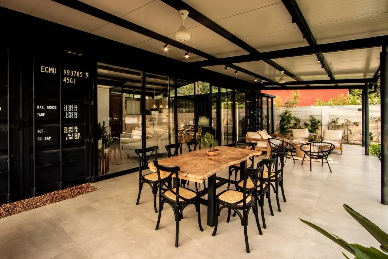 Galeria de lujo de casa en contenedor negro con marcas navieras a la vista, comedor de madera viva, sillones tejidos y paredes de vidrio que muestran el living. La historia contenedor a lujo en una toma.