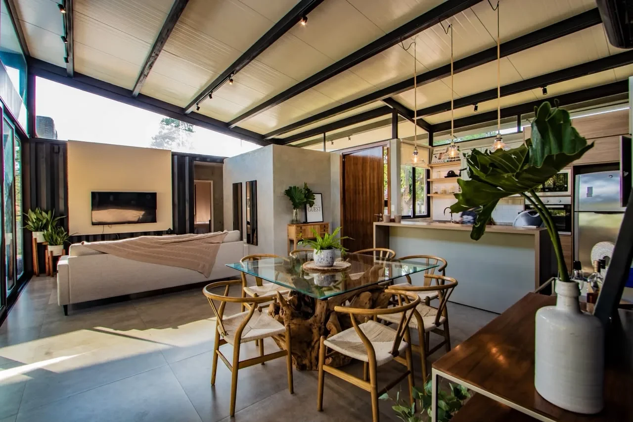 Espacio integrado living-comedor-cocina de la casa de lujo en contenedor, con sofa gris, mesa de vidrio sobre tronco, sillas wishbone y paredes de vidrio al jardin.