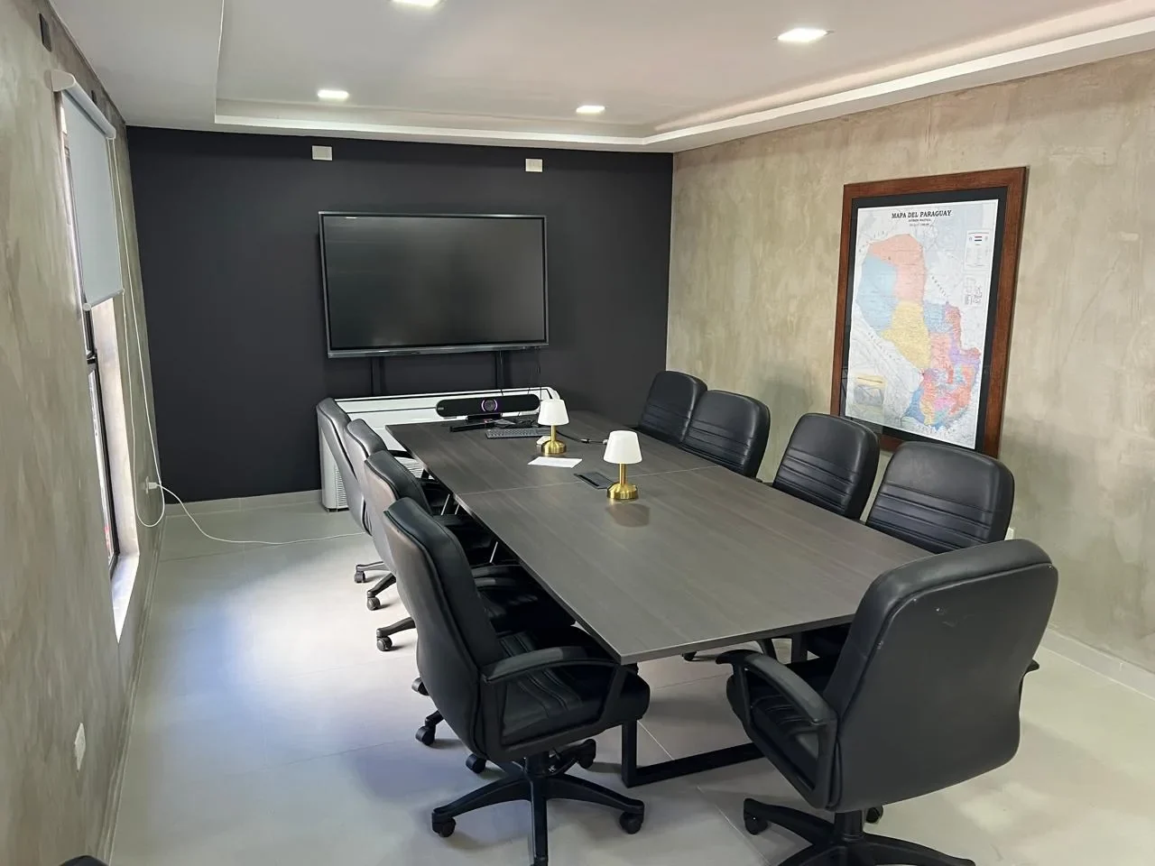 Sala de reuniones en contenedor con mesa de directorio, lamparas de bronce, pantalla y mapa del Paraguay sobre microcemento.