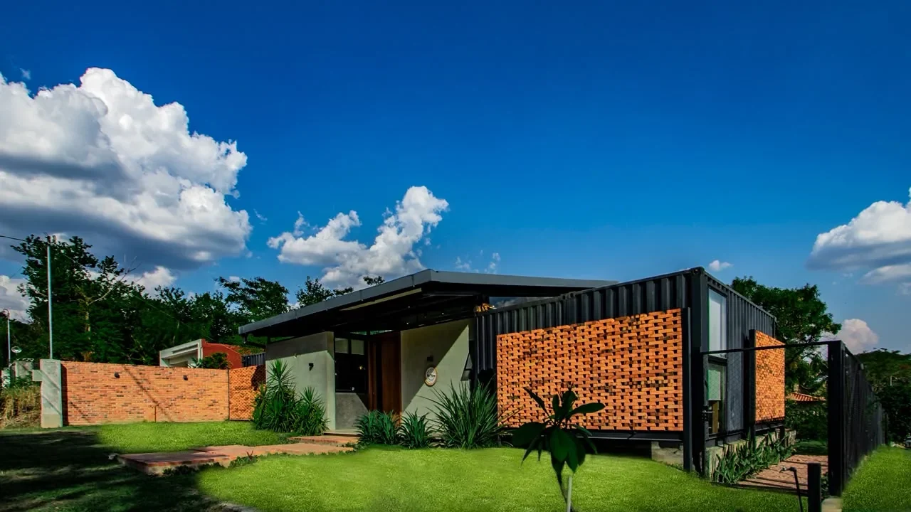 Casa de contenedor negro con celosias de ladrillo rojo calado y entrada de microcemento con puerta de madera, cesped y cielo dramatico. Arquitectura contemporanea.