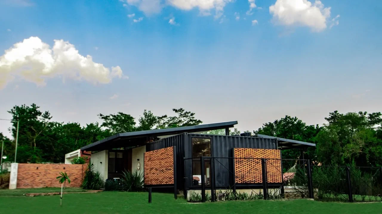 Casa de contenedor negro con celosias de ladrillo rojo y techo mariposa, sobre amplio jardin con hamaca. Arquitectura contemporanea en Paraguay.