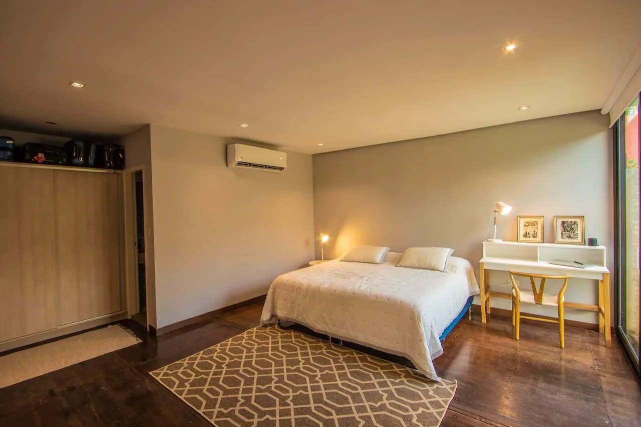 Dormitorio editorial en casa de contenedor con cama de acolchado capitone, escritorio con silla wishbone y pared de vidrio hacia el patio con muro de ladrillo y enredadera.
