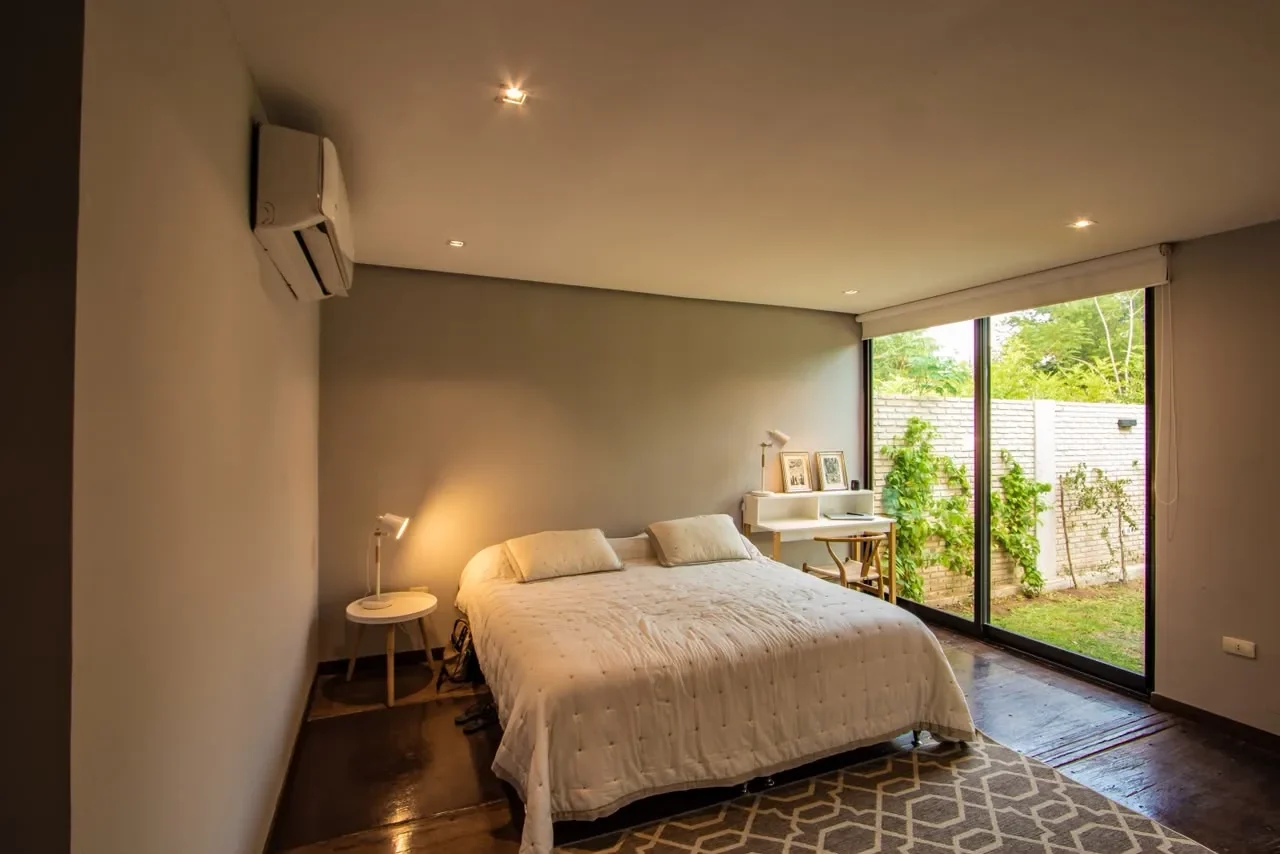 Dormitorio de diseno en casa de contenedor con pared de vidrio al patio de ladrillo con enredadera, cama con acolchado capitone, escritorio con silla wishbone y alfombra geometrica.