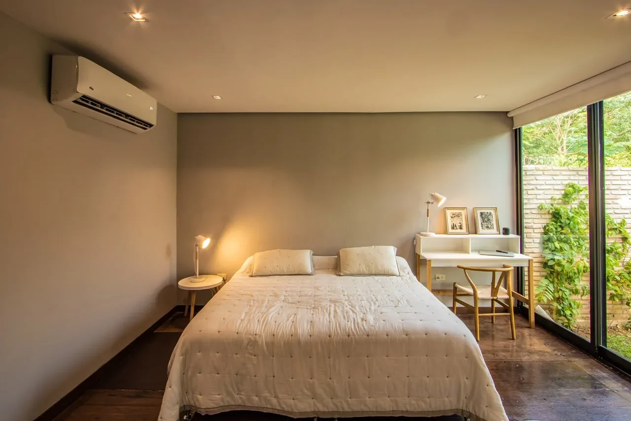 Dormitorio de diseno en casa de contenedor con pared de vidrio hacia un patio con muro de ladrillo y enredadera, acolchado capitone y escritorio con silla wishbone.