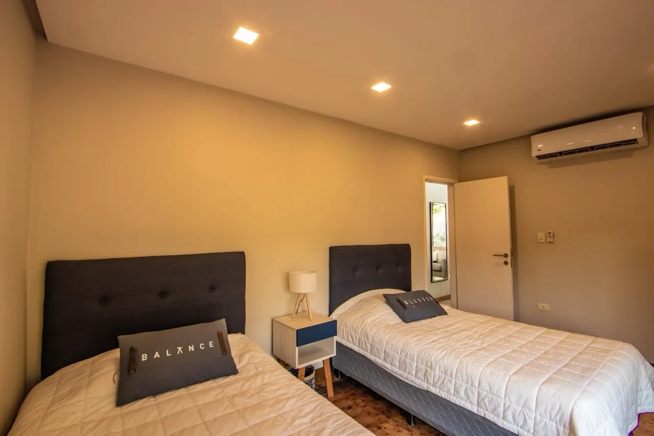 Dormitorio de huespedes editorial en casa de contenedor con dos camas, cabeceras tapizadas azules y almohadones 'Balance', salida a la terraza. Estilo posada.
