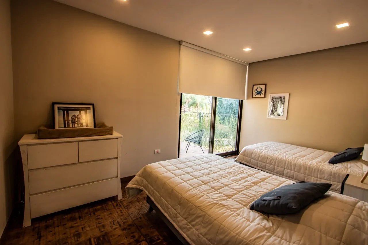 Dormitorio de huespedes editorial en casa de contenedor con dos camas individuales, comoda blanca, cuadros y pared de vidrio a la terraza, piso de madera.