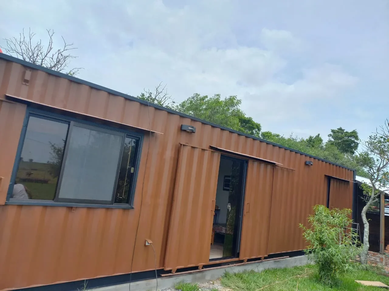 Casa de contenedor corten marron con puerta corrediza de vidrio abierta y ventana, en entorno rural con campo verde.
