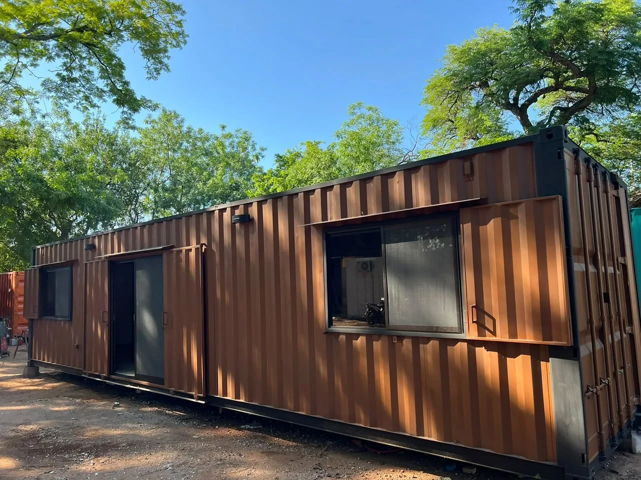 Modulo de contenedor maritimo pintado en corten marron cobrizo, con ventana de aluminio negro y puerta corrediza de vidrio, entre arboles.