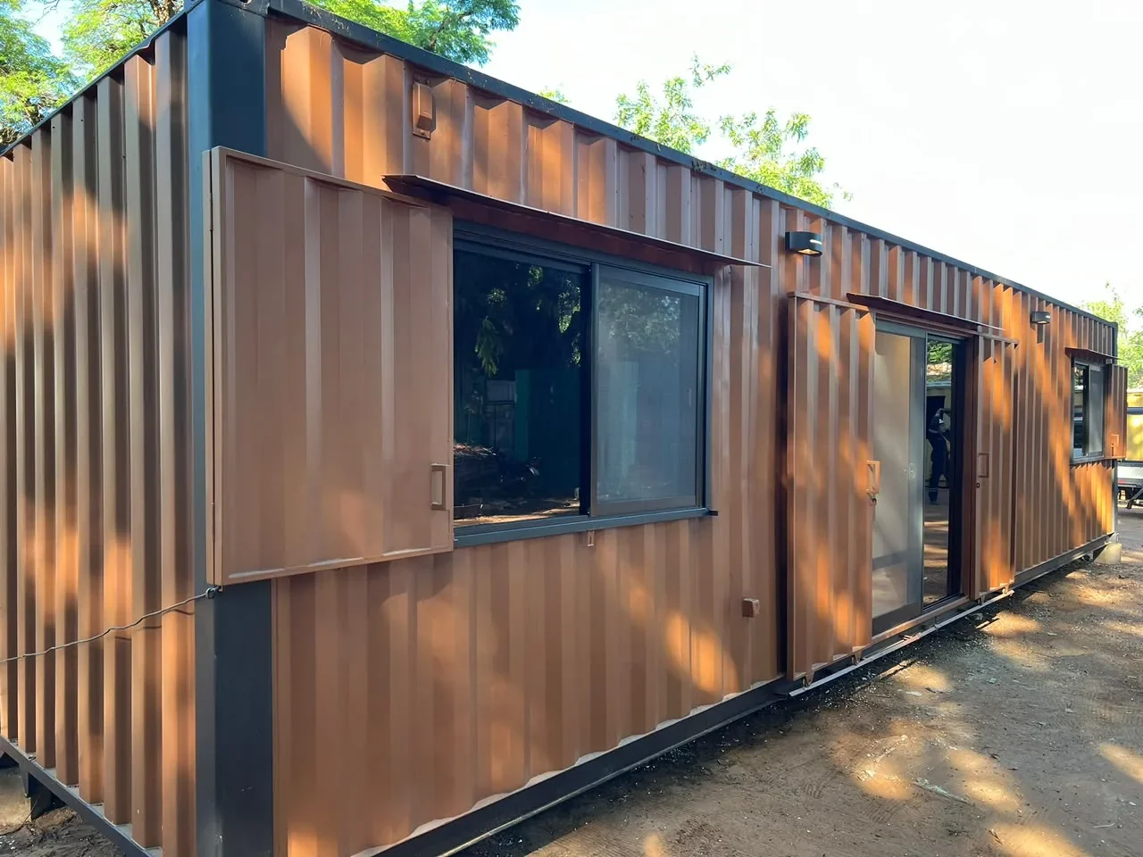 Modulo de contenedor corten marron con postigos rebatibles abiertos y puerta corrediza de vidrio, en predio de tierra.