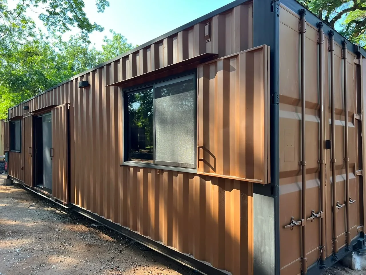 Lateral del modulo de contenedor corten marron con ventana corrediza de marco negro, entre arboles.